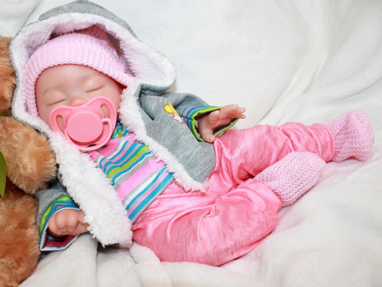 Sleeping reborn silicone baby doll wearing pink knit hat, striped shirt, and pink pants, lifelike 13-inch newborn doll on white blanket