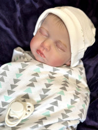 Thumbnail for A baby doll rests on a soft blanket, showcasing its delicate features and clothing.