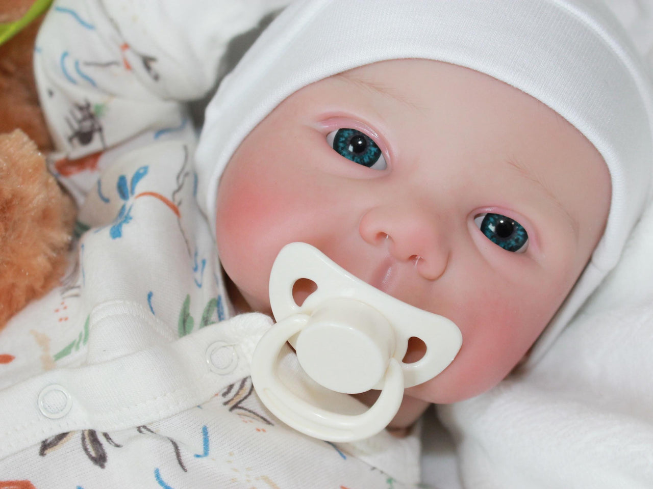 A baby doll positioned on a clean white sheet, with a pacifier, highlighting its charming design and soft textures.