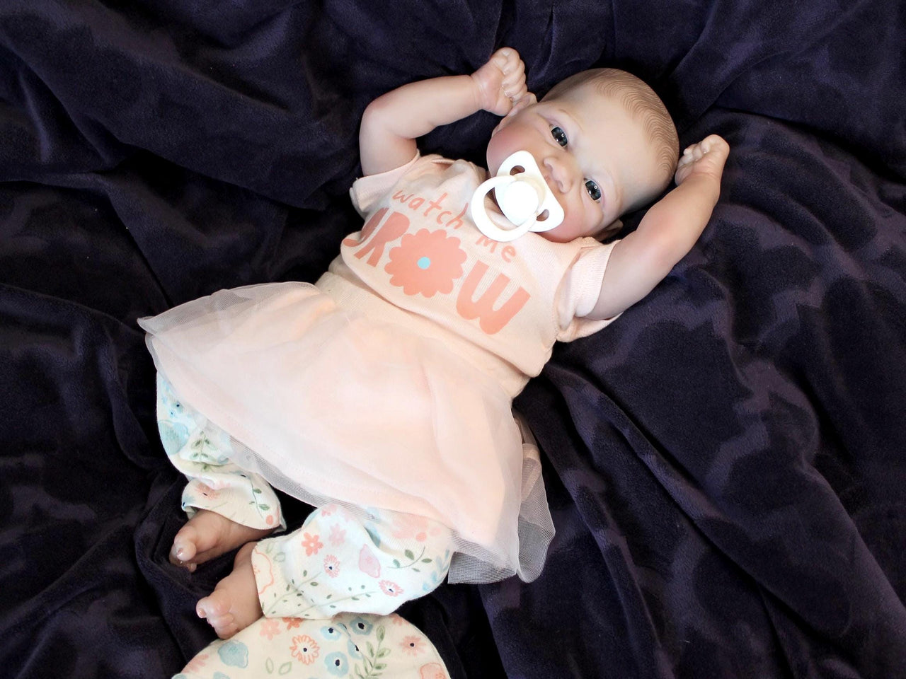 A baby doll with a pacifier lies on a cozy blanket, highlighting its charming design and soothing presence.