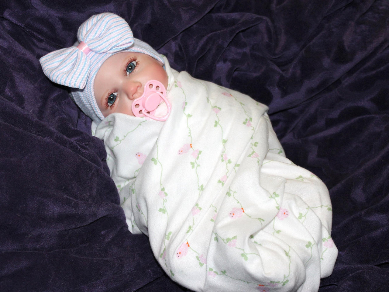 Lifelike reborn baby doll with blue eyes wearing a pink floral outfit and striped bow hat, realistic weighted newborn therapy doll