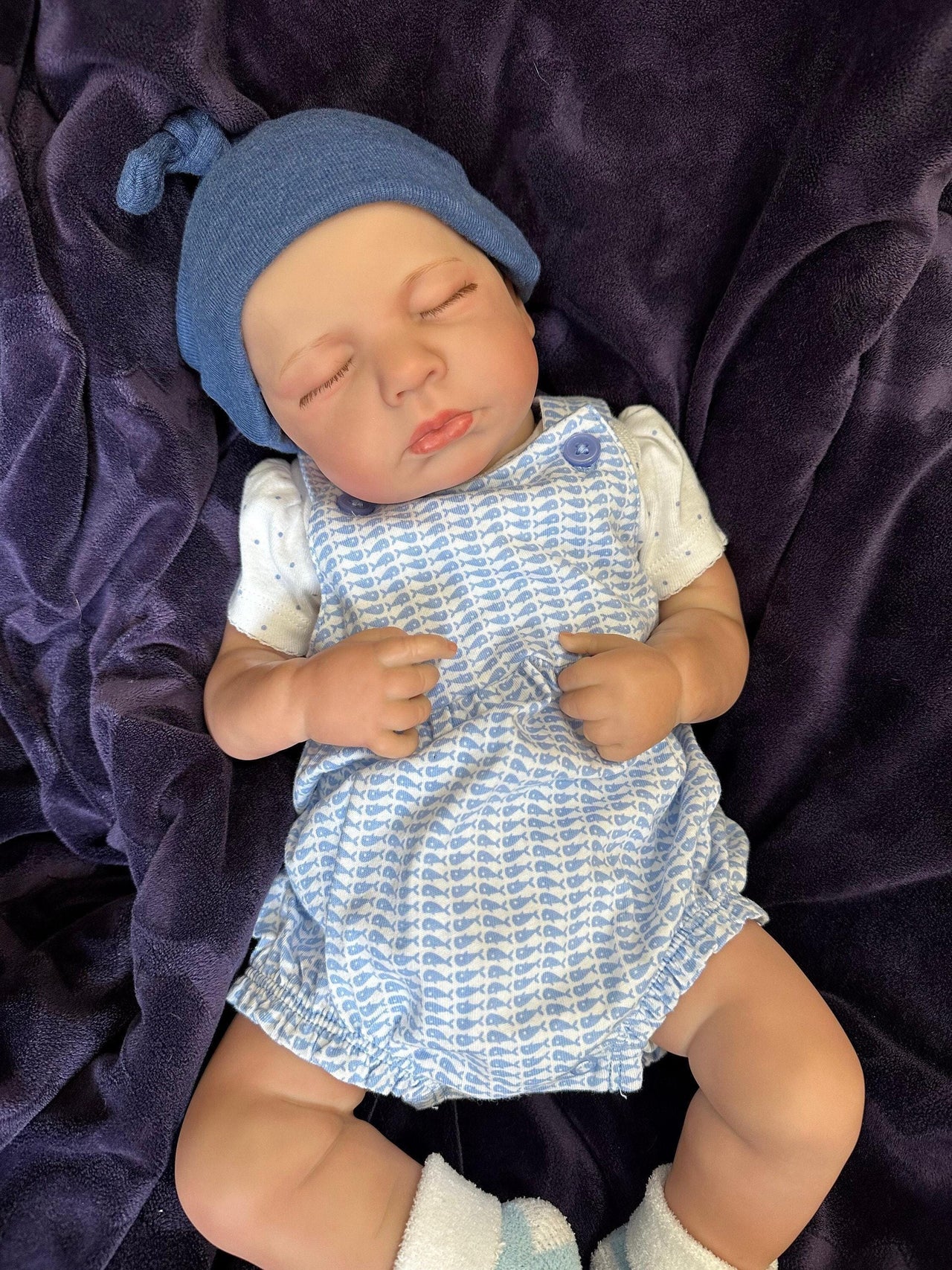 Sleeping reborn baby doll wearing blue knit hat, blue checkered romper, and white socks on purple blanket – realistic handmade newborn doll