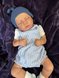 Thumbnail for Sleeping reborn baby doll wearing blue knit hat, blue checkered romper, and white socks on purple blanket – realistic handmade newborn doll