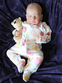 Thumbnail for Sleeping reborn baby doll in fruit-print pajamas, holding a small tan teddy bear, realistic handmade newborn doll on purple blanket