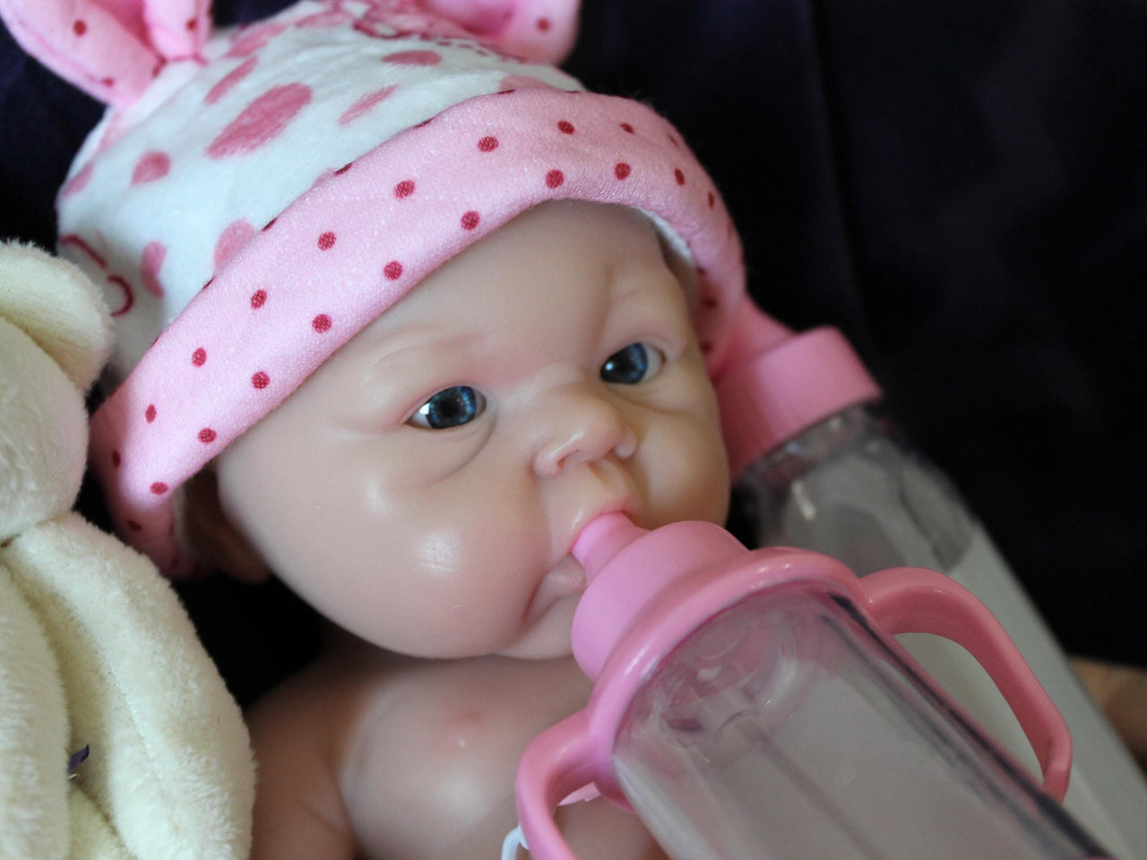 Lifelike reborn baby doll with blue eyes wearing pink polka dot outfit and bunny ear hat, realistic handmade newborn doll on blanket
