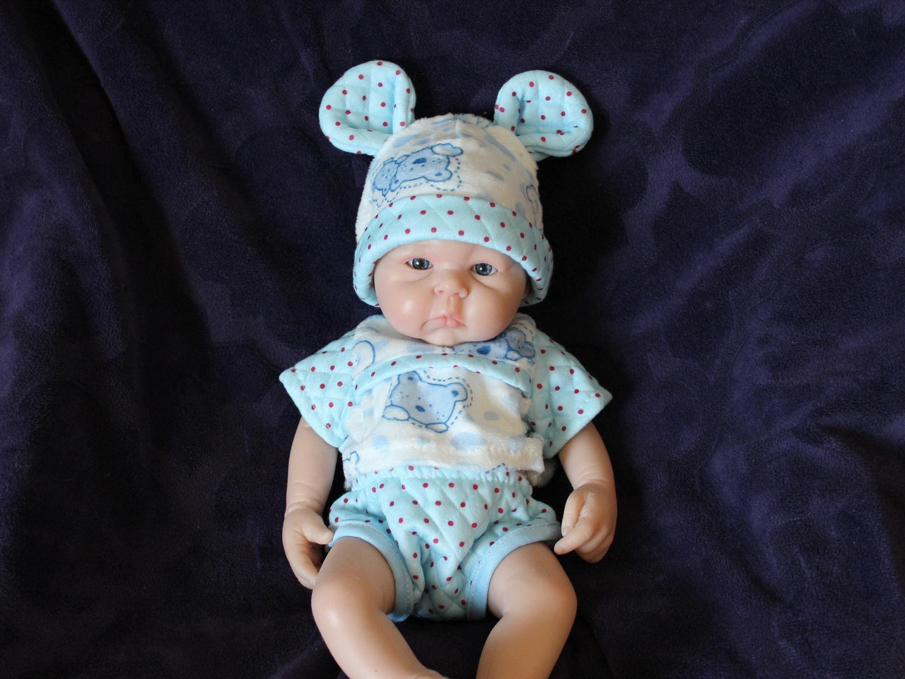 Lifelike reborn baby doll with blue eyes wearing blue bear-print outfit and matching bunny ear hat, realistic handmade newborn doll