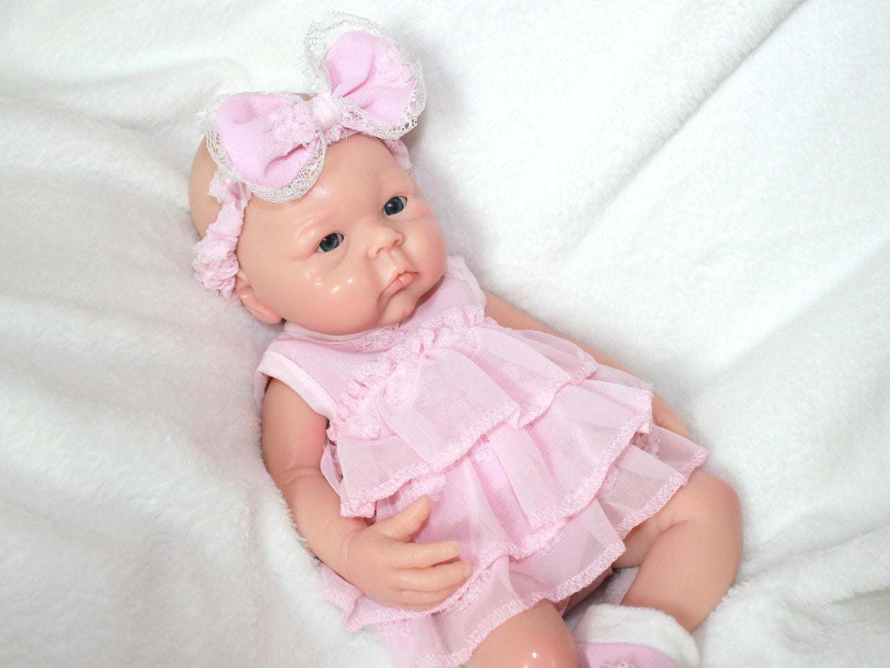 A baby doll dressed in a pink dress and matching headband, sitting against a soft background.