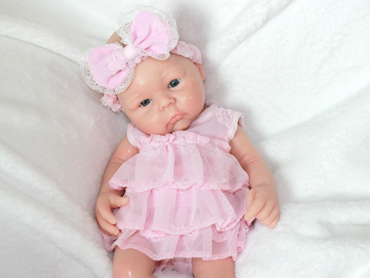 A baby doll dressed in a pink dress and matching headband, sitting against a soft background.