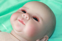 Thumbnail for A baby doll resting on a green sheet, showcasing its soft features and delicate clothing.