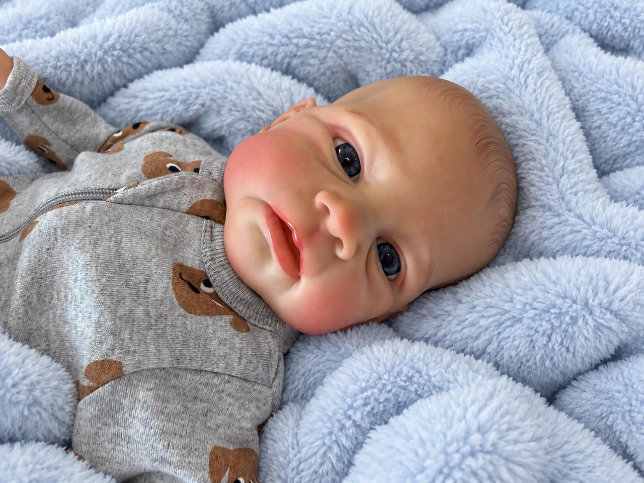 Realistic reborn baby doll featuring lifelike newborn details, delicate facial features, realistic skin texture, hand-painted details, and beautiful expressive eyes. Photographed resting naturally on a soft, cozy baby blanket to showcase the doll's realistic appearance and craftsmanship.