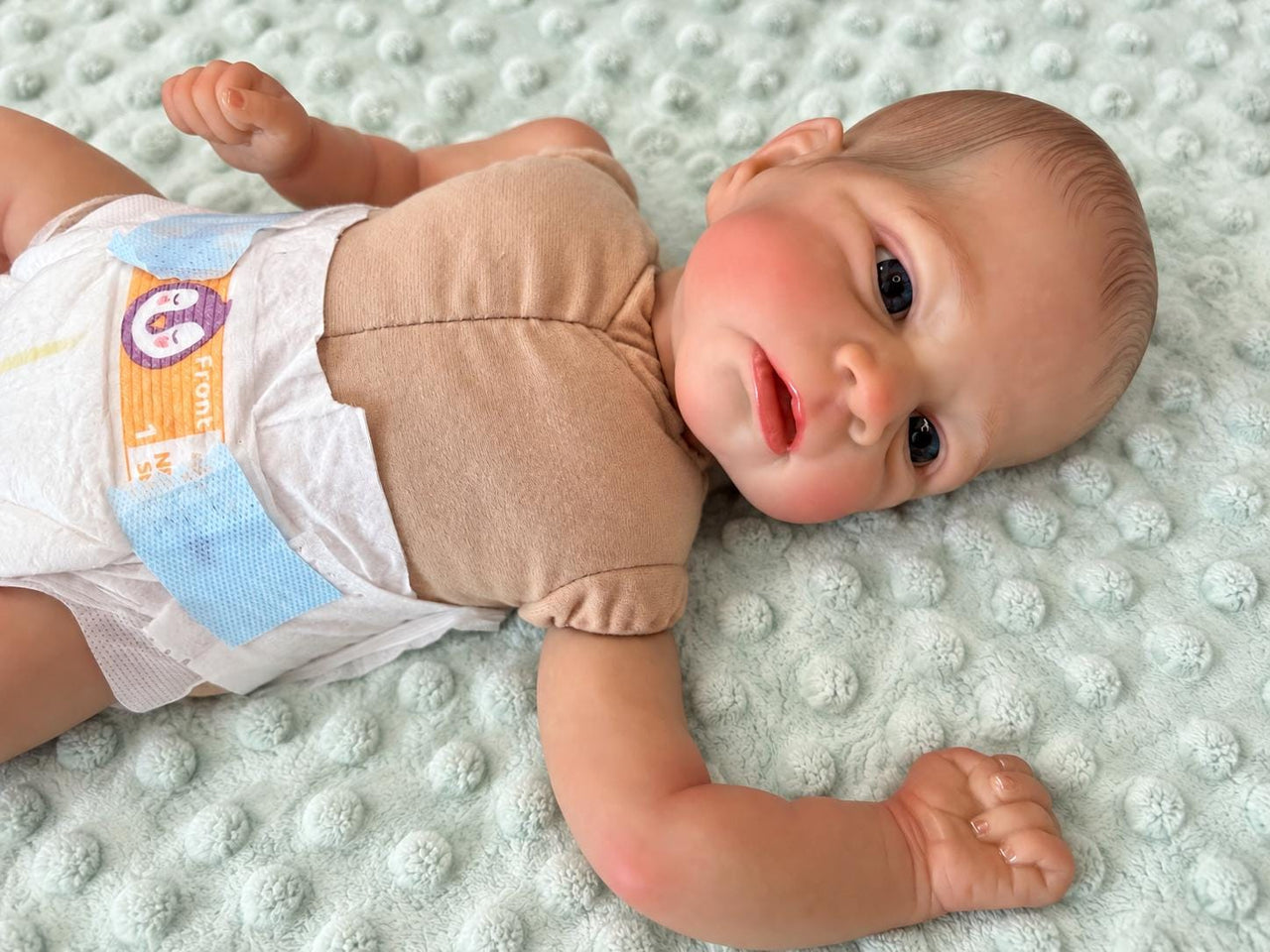 Realistic reborn baby doll featuring lifelike newborn details, delicate facial features, realistic skin texture, hand-painted details, and beautiful expressive eyes. Photographed resting naturally on a soft, cozy baby blanket to showcase the doll's realistic appearance and craftsmanship.