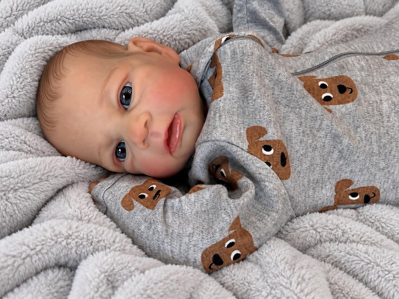 Realistic reborn baby doll featuring lifelike newborn details, delicate facial features, realistic skin texture, hand-painted details, and beautiful expressive eyes. Photographed resting naturally on a soft, cozy baby blanket to showcase the doll's realistic appearance and craftsmanship.