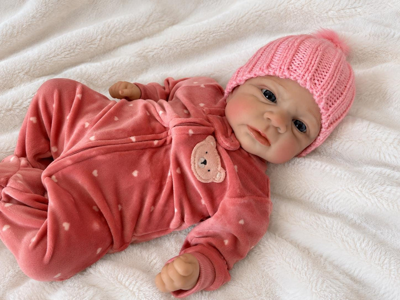 Realistic reborn baby doll featuring lifelike newborn details, delicate facial features, realistic skin texture, hand-painted details, and beautiful expressive eyes. Photographed resting naturally on a soft, cozy baby blanket to showcase the doll's realistic appearance and craftsmanship.