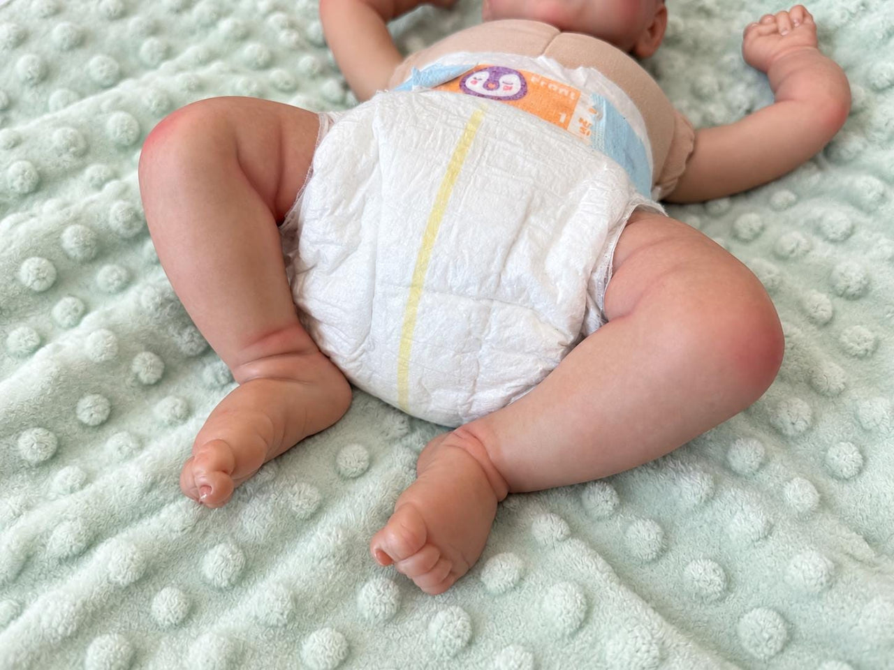 Realistic reborn baby doll featuring lifelike newborn details, delicate facial features, realistic skin texture, hand-painted details, and beautiful expressive eyes. Photographed resting naturally on a soft, cozy baby blanket to showcase the doll's realistic appearance and craftsmanship.