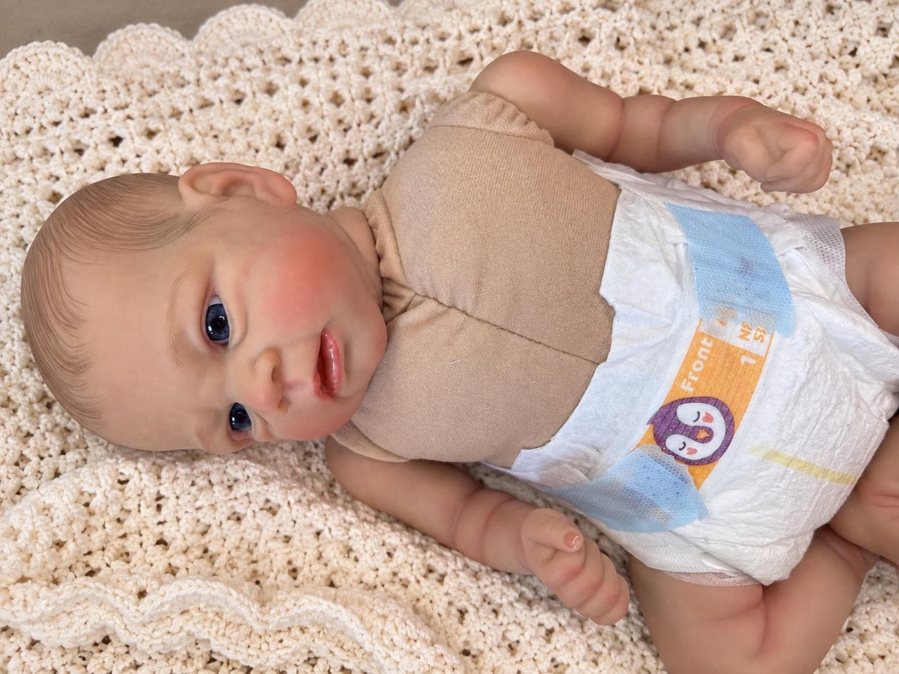 Realistic reborn baby doll featuring lifelike newborn details, delicate facial features, realistic skin texture, hand-painted details, and beautiful expressive eyes. Photographed resting naturally on a soft, cozy baby blanket to showcase the doll's realistic appearance and craftsmanship.